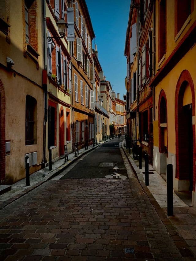 Une rue pavée étroite et vide, bordée de bâtiments historiques colorés aux volets clos, sous un ciel bleu clair.