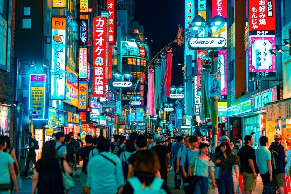 A crowd of people walks down a busy street at night, surrounded by tall buildings covered in bright, colorful Japanese neon signs.