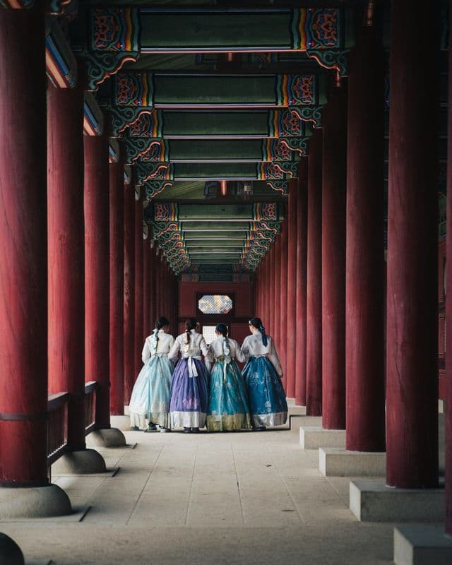 Four women wearing colorful traditional hanbok dresses walk down a covered corridor lined with red pillars.
