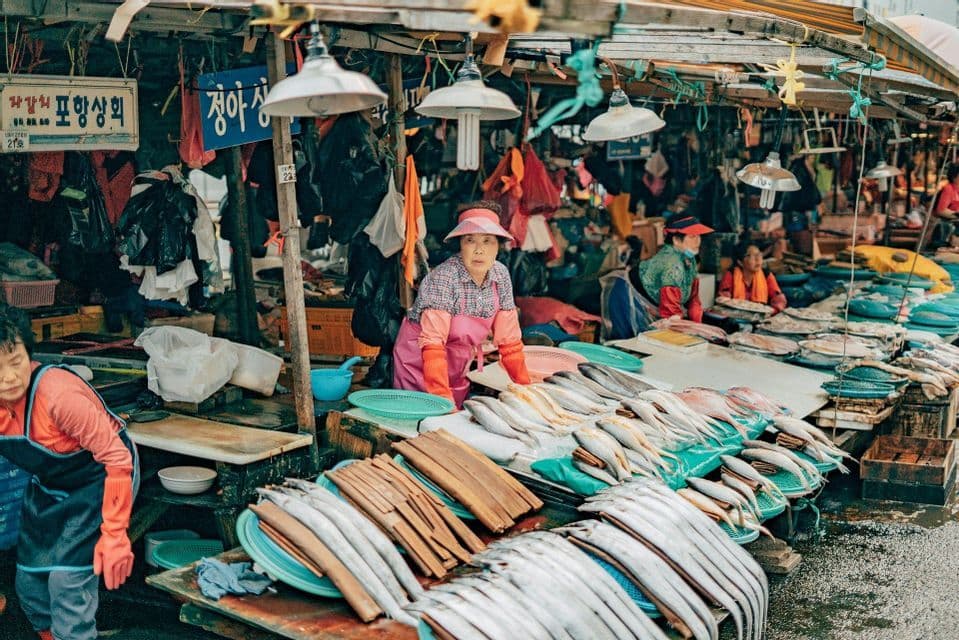 A woman in a pink apron stands at a stall in a busy outdoor fish market, with fresh fish displayed on tables.