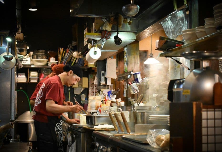 A chef in a red shirt and black bandana cooks with chopsticks in a busy, steamy restaurant kitchen filled with equipment.