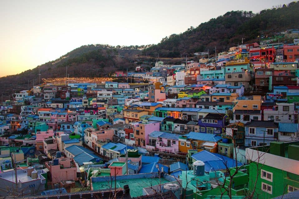 A dense hillside village of colorful houses with bright blue, pink, and green roofs, viewed at sunset.