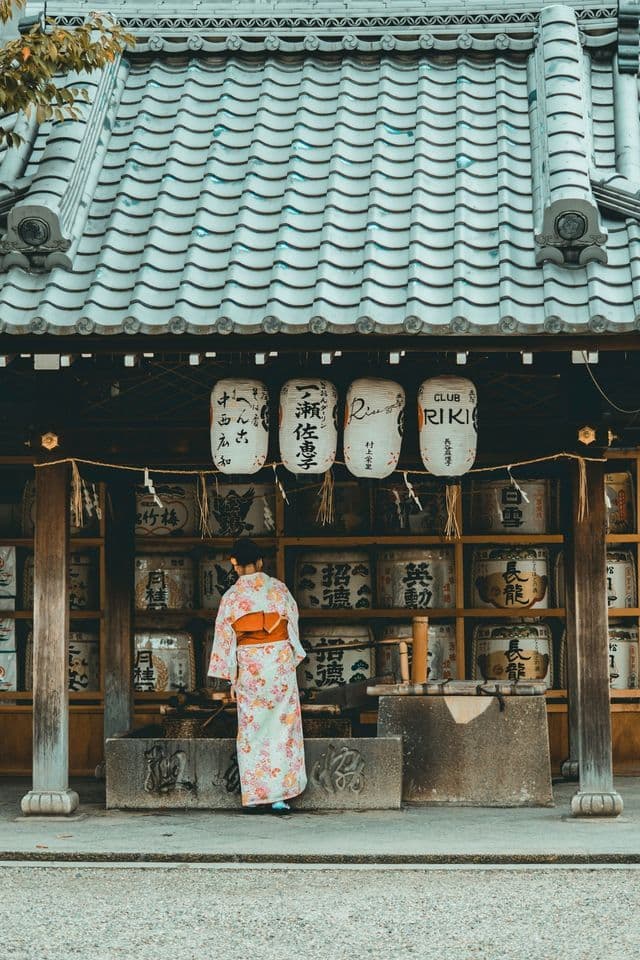 A woman from behind in a floral kimono stands at a traditional Japanese building with paper lanterns and sake barrels.