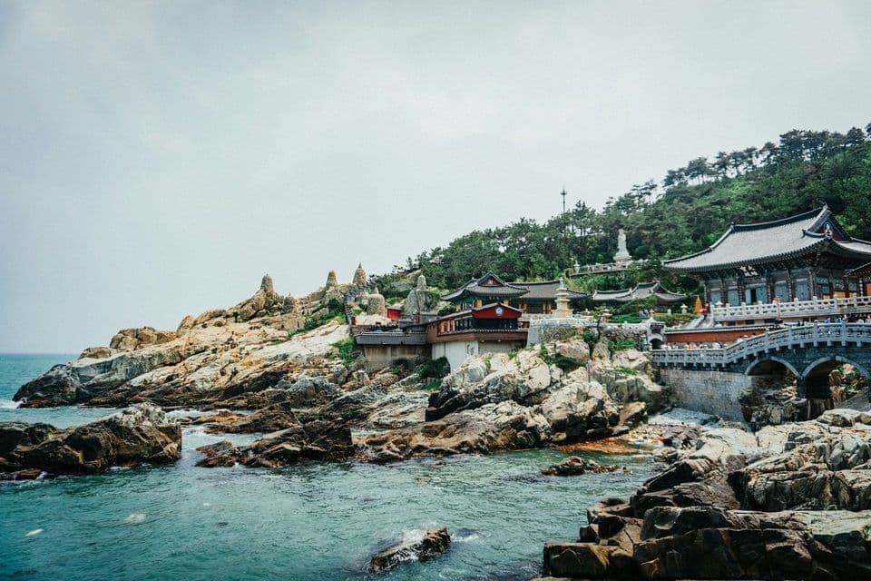 A traditional Asian temple complex built on a rocky coastline, with a forested hill behind it and the sea in front.