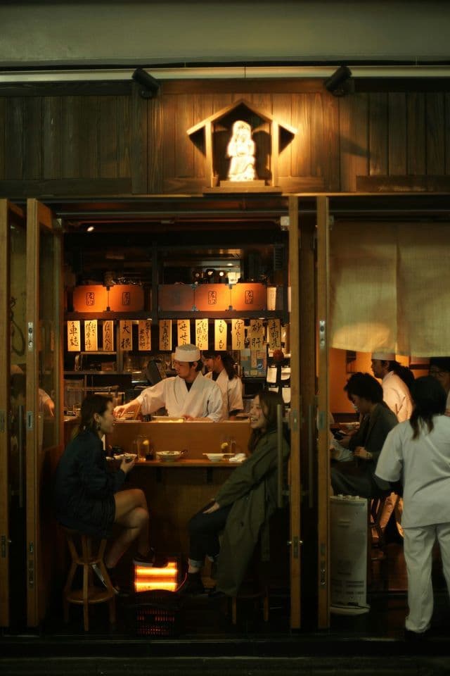 Two women eat and talk at the counter of a traditional Japanese restaurant at night, viewed from the street.