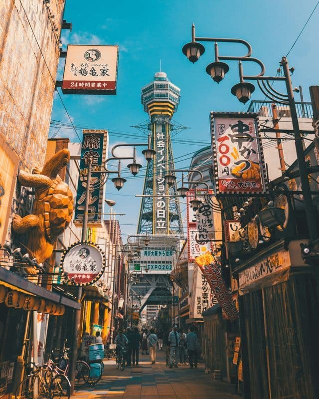 A low-angle view of a busy city street lined with buildings with Japanese signs, leading to a tall observation tower under a blue sky.