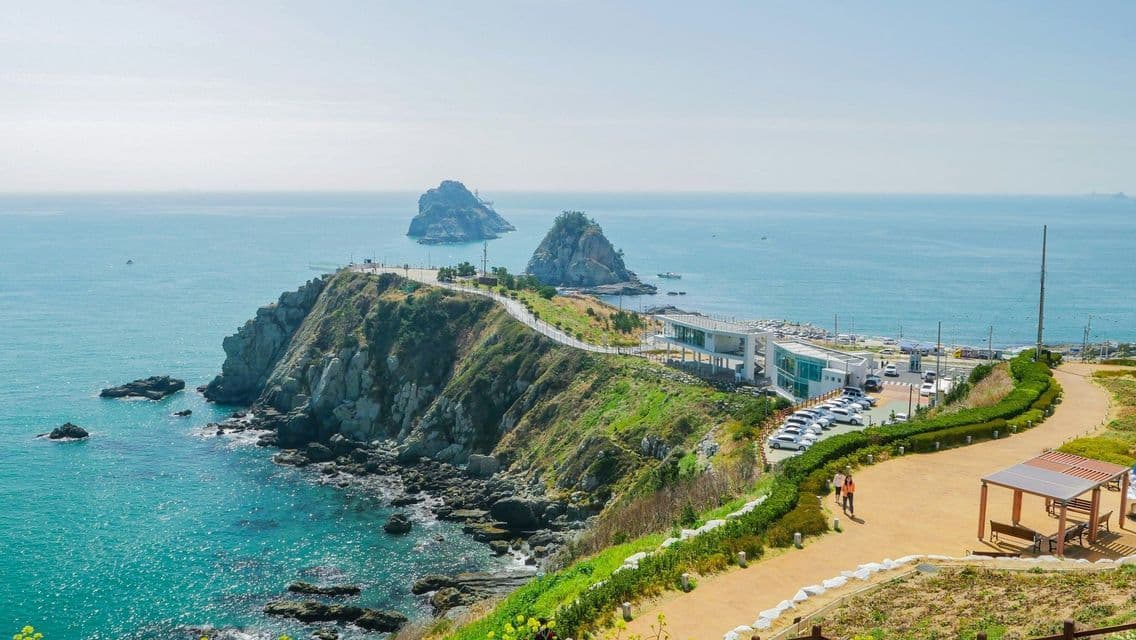 A high-angle view of a verdant, rocky coastline with a winding path, overlooking a turquoise sea with two prominent islets.