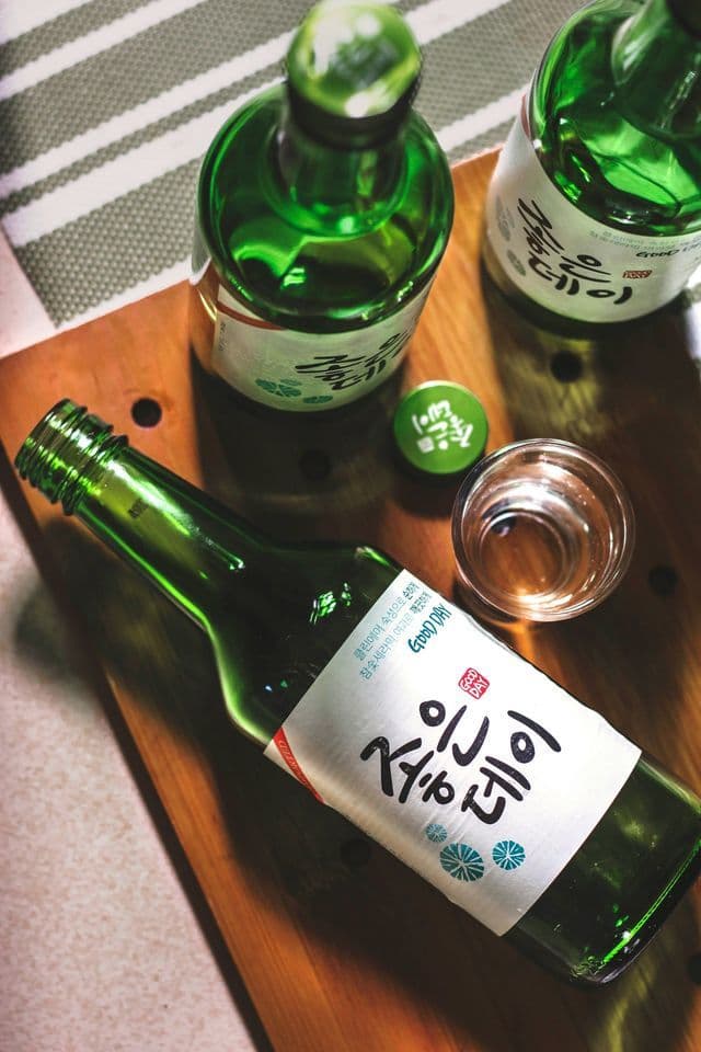 An overhead shot of green soju bottles, a bottle cap, and a filled shot glass on a wooden board.