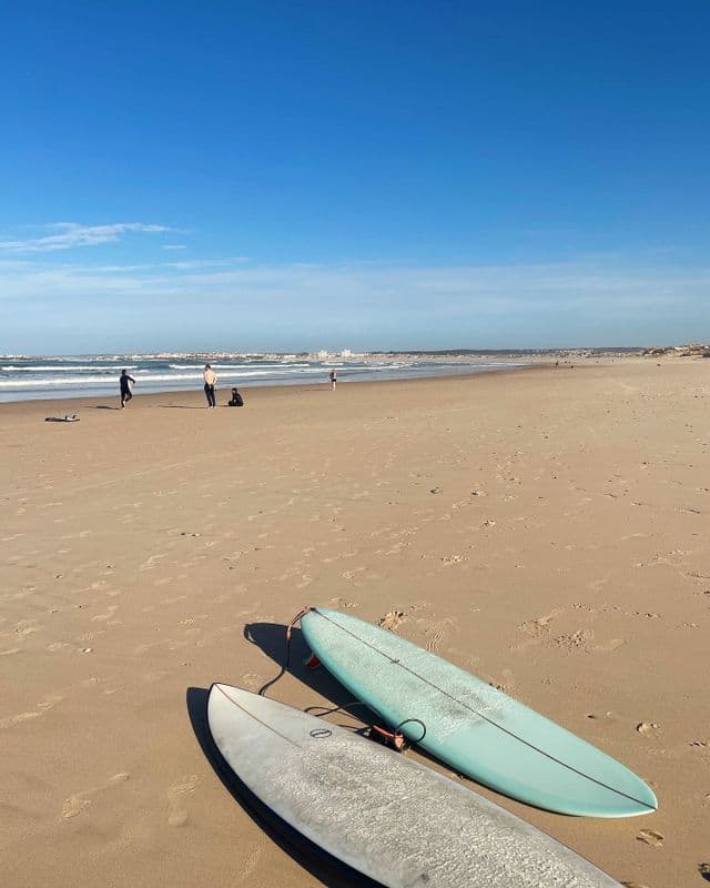 Two surfboards rest on a sandy beach with people and waves in the distance under a clear blue sky.