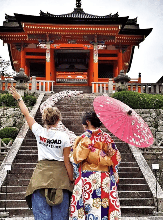 A WeRoad group leader stands with a woman in a kimono and parasol on stairs leading to a traditional Japanese temple.