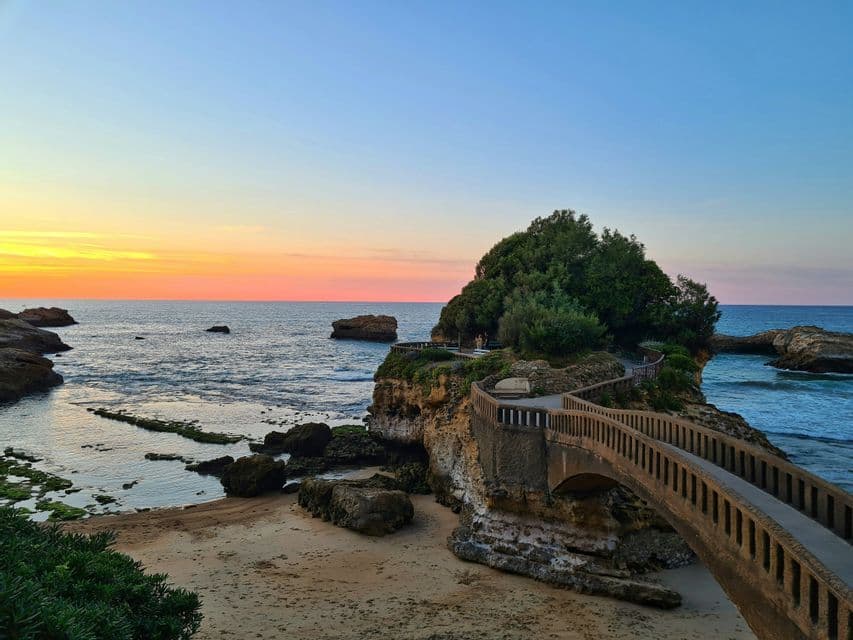 Un pont de pierre s'étend d'une plage de sable vers un petit îlot rocheux arboré, sous un coucher de soleil coloré sur l'océan.