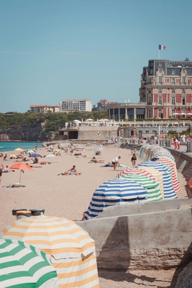 Una spiaggia assolata, affollata di bagnanti e ombrelloni a righe colorate, con un grande edificio che espone una bandiera francese sullo sfondo.