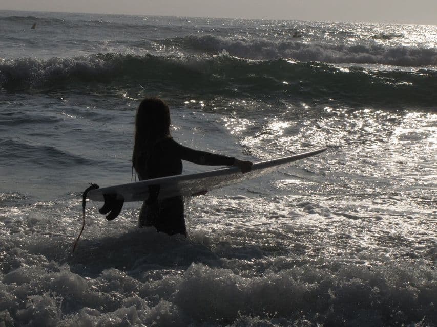 Un surfeur en combinaison, dont la silhouette se découpe, tient une planche de surf en pénétrant dans l'eau agitée de l'océan, scintillante sous le soleil.