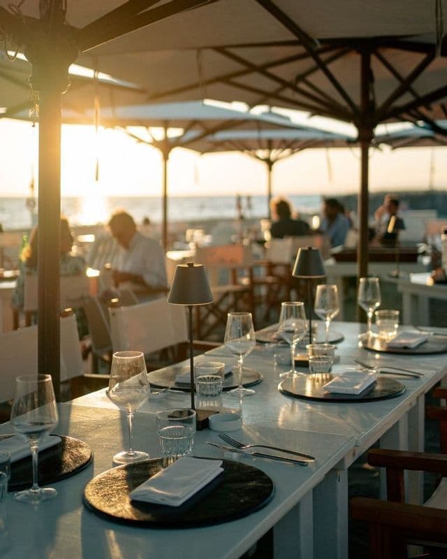 Un lungo tavolo da pranzo apparecchiato con bicchieri e luci in un ristorante sulla spiaggia all'aperto durante un tramonto dorato.