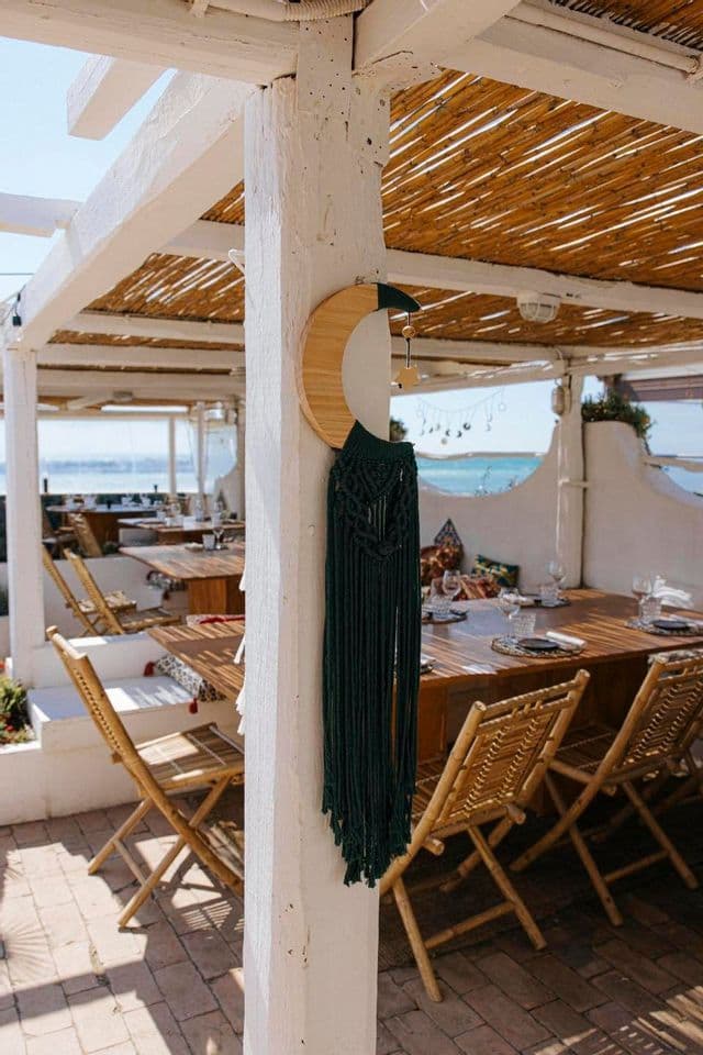 Una decorazione macramè a mezzaluna pende su un pilastro bianco in un ristorante all'aperto sul mare, con tavoli in legno e un tetto di paglia.