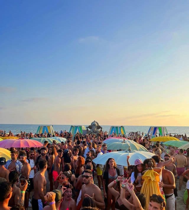 Una grande folla di persone che fanno festa su una spiaggia sabbiosa al tramonto, con ombrelloni colorati e un palco di fronte all'oceano.