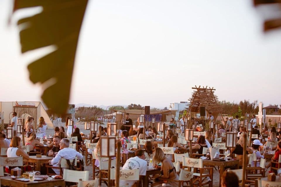 Una folla di persone che socializza a tavoli di legno in un ristorante sulla spiaggia all'aperto, al tramonto, con lanterne sui tavoli.