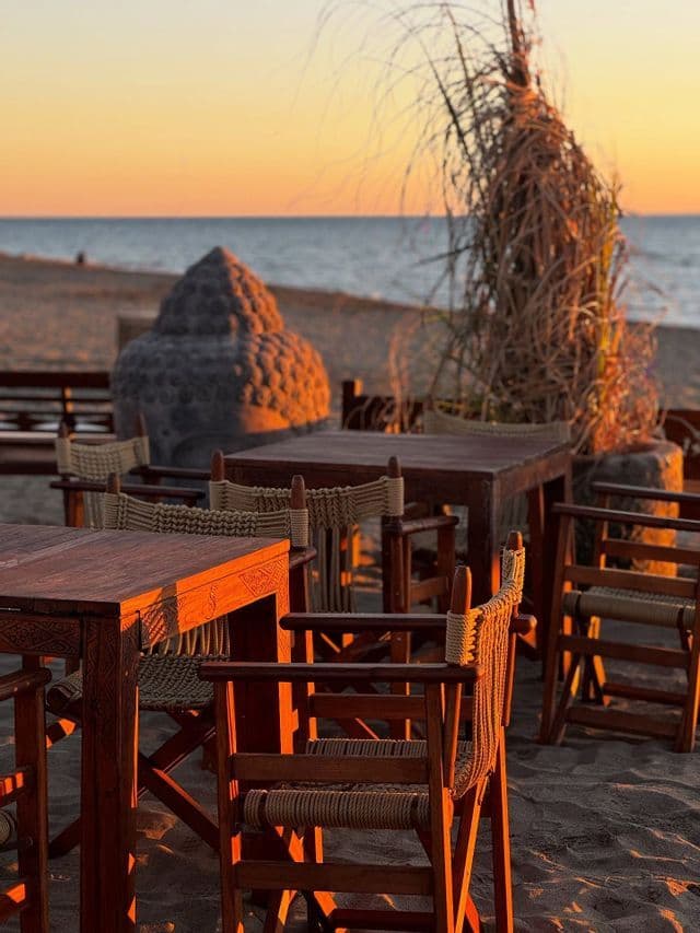Tavoli in legno e sedie in vimini su una spiaggia di sabbia affacciata sull'oceano al tramonto, con una grande testa di Buddha in pietra lì vicino.