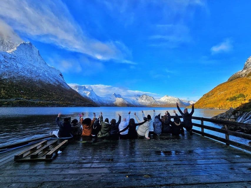 Un grupo de viaje de WeRoad está en un muelle de madera con las manos levantadas, contemplando montañas nevadas al otro lado de un lago bajo un cielo azul.