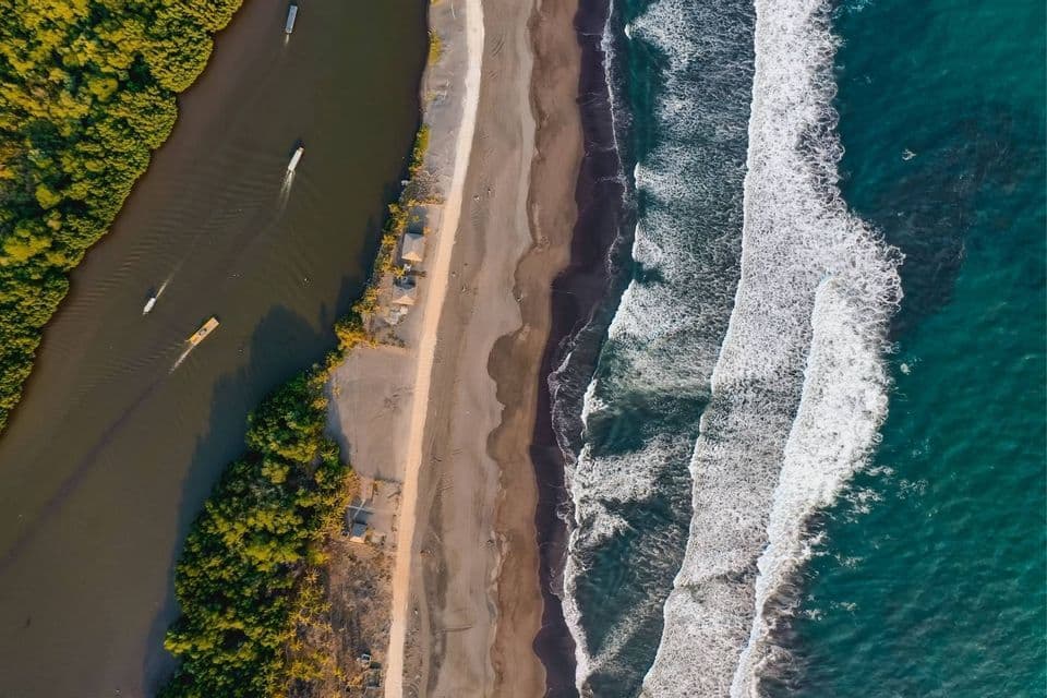 Eine Luftaufnahme zeigt einen ruhigen Fluss mit Booten, der parallel zu einem Sandstrand und dem türkisfarbenen Meer mit brechenden Wellen verläuft.