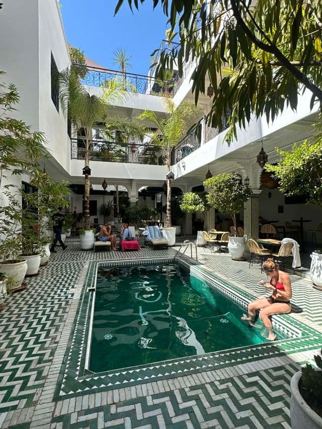 Un voyage de groupe WeRoad se détendant au bord d'une piscine dans la cour ensoleillée et carrelée d'un riad avec des plantes luxuriantes.