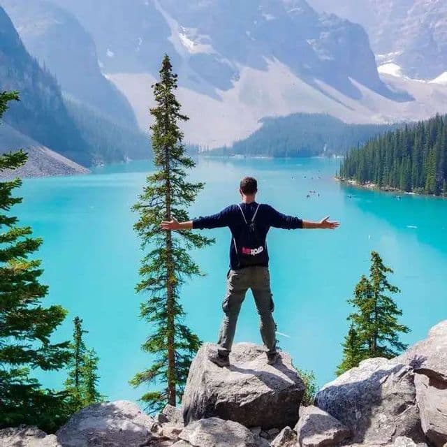 Un uomo con uno zaino WeRoad si trova su una roccia con le braccia distese, ammirando un lago turchese e le montagne.