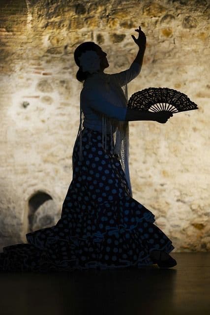 Eine Silhouette einer Flamenco-Tänzerin in einem gepunkteten Kleid, die mit einem schwarzen Spitzenfächer vor einer beleuchteten Steinwand posiert.