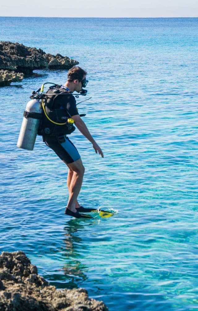 Un homme en tenue de plongée complète s'apprête à entrer dans les eaux bleues et claires depuis un rivage rocheux.