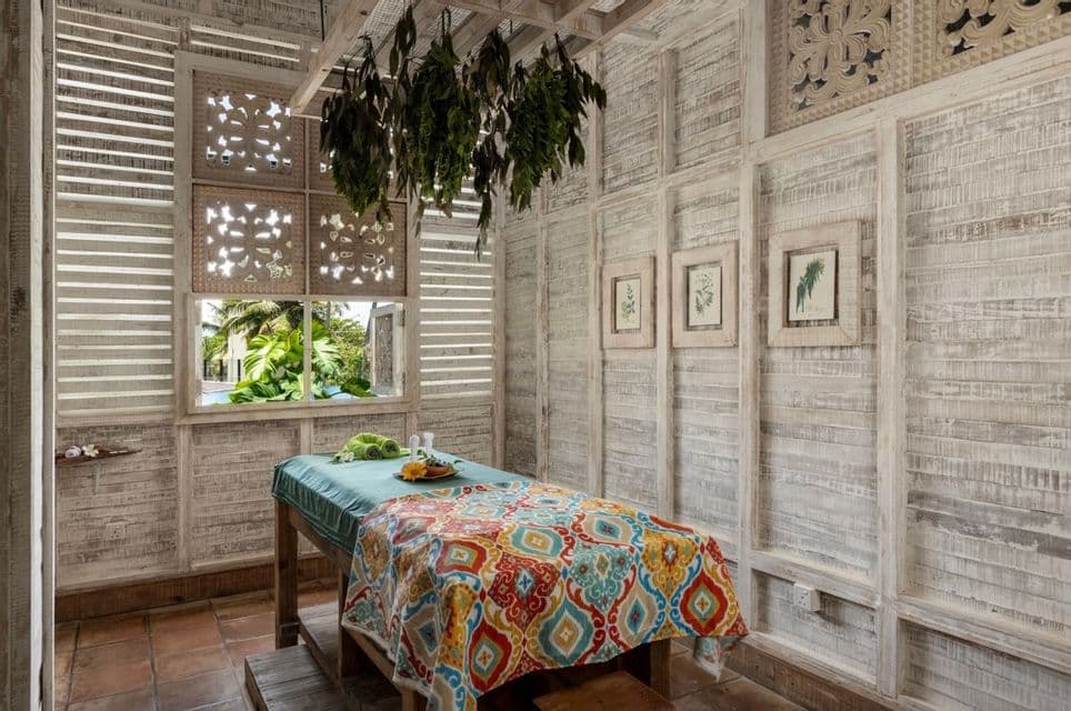 Une table de massage avec une couverture colorée dans une pièce rustique aux murs blanchis à la chaux, des herbes suspendues et une fenêtre donnant sur la verdure.