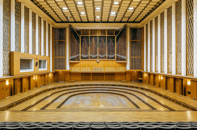 Un'ampia veduta di una sala da concerto vuota, rivestita in legno, con un grande organo a canne sopra un palco a gradoni.