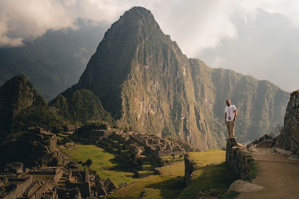 Un hombre se encuentra en un saliente de piedra con vistas a ruinas antiguas enclavadas entre montañas verdes bajo un cielo parcialmente nublado.