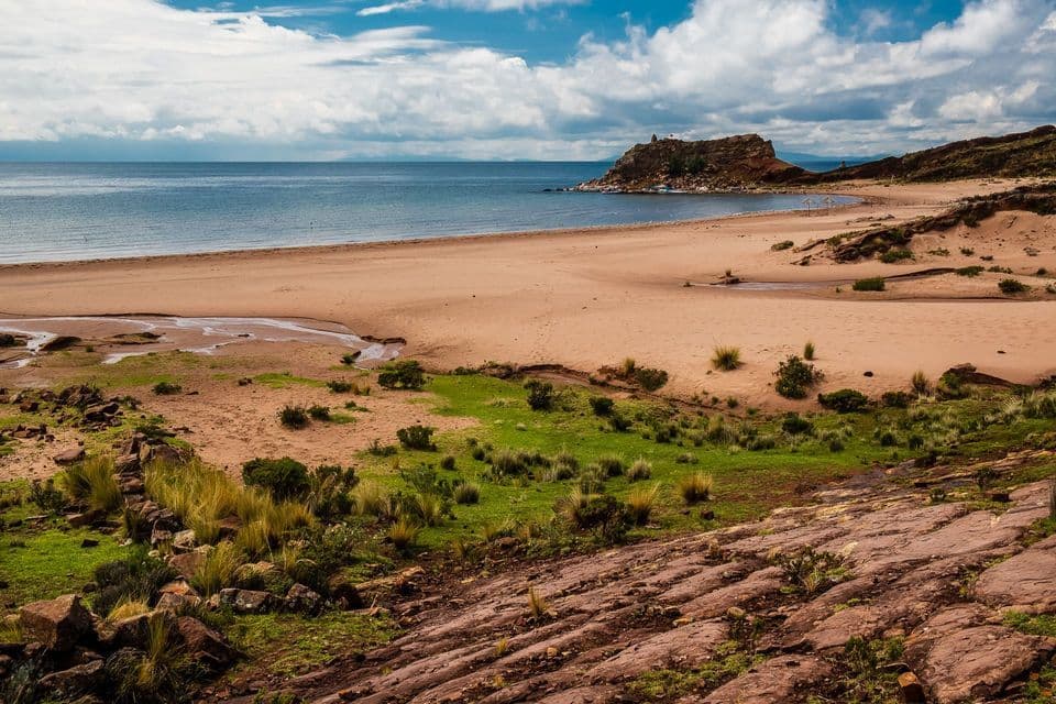 A wide, sandy beach beside a calm body of water, with green grassy patches and a rocky headland under a partly cloudy sky.