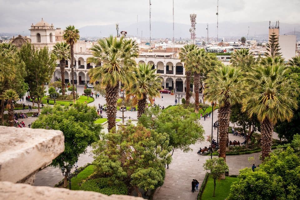 An elevated view of a bustling city plaza filled with palm trees, green spaces, and people walking among historic buildings under a cloudy sky.