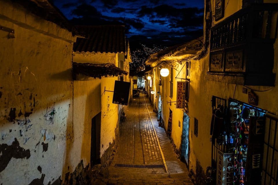 Un callejón empedrado, estrecho y cuesta abajo, iluminado por farolas cálidas bajo un cielo nocturno oscuro y nublado.