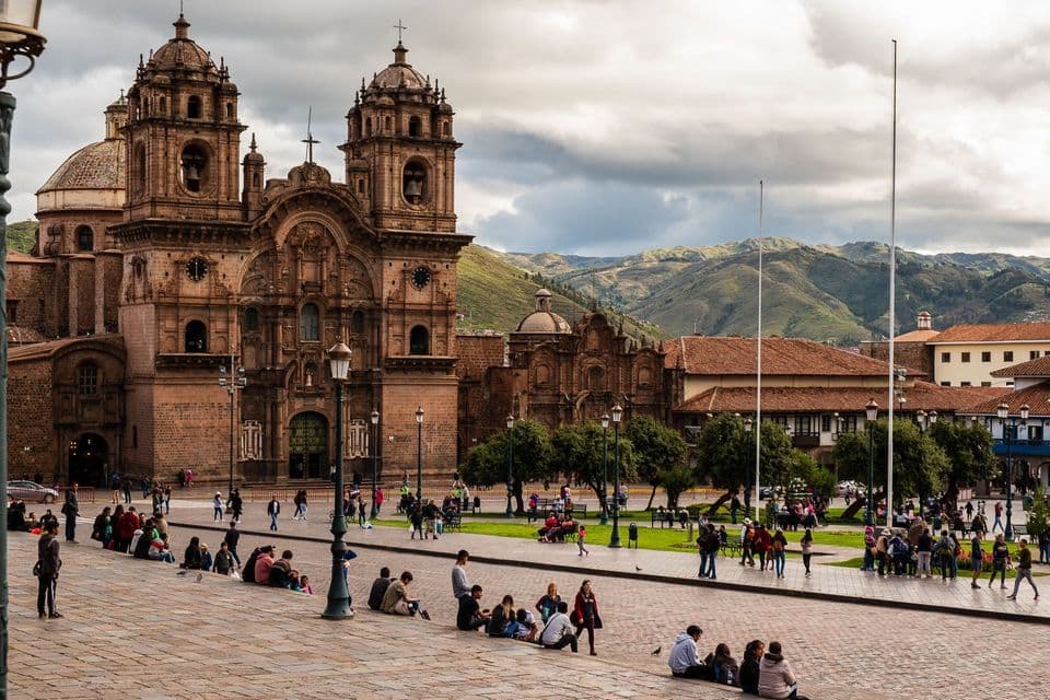 Una multitud de personas se reúne en una gran plaza de piedra frente a una ornamentada catedral histórica, con montañas verdes elevándose al fondo.