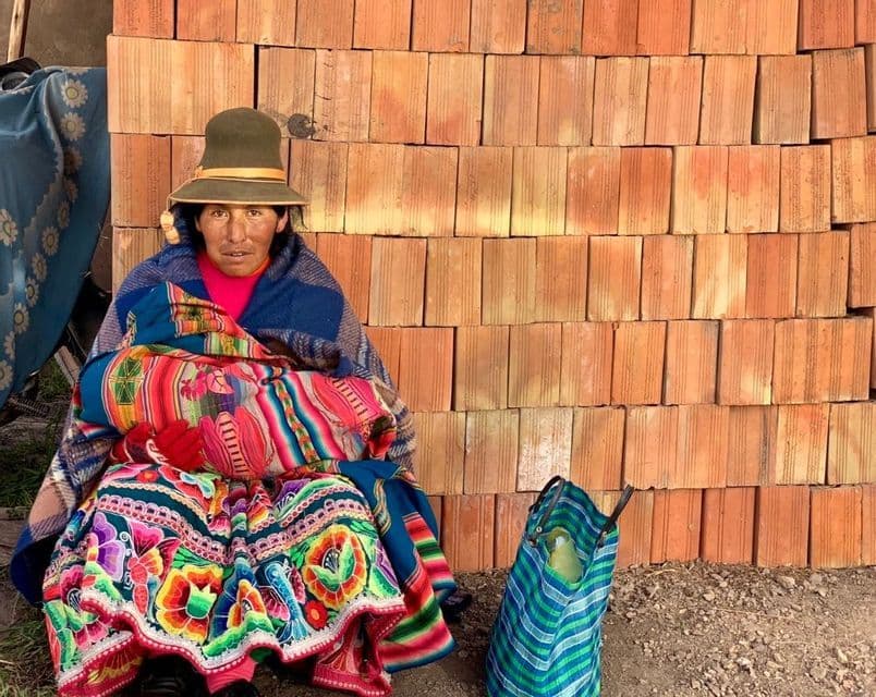 Eine Person mit Hut und farbenfroher Tracht sitzt auf dem Boden vor einer Wand aus gestapelten orangefarbenen Ziegeln.