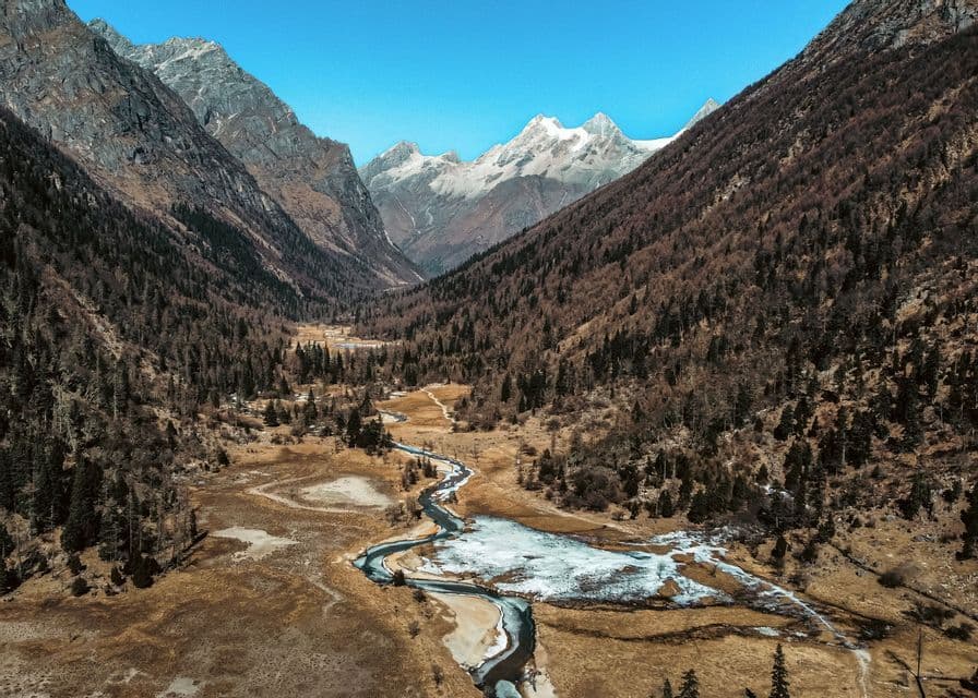 An aerial view of a partially frozen river winding through a vast valley between steep, forested mountains with snow-capped peaks in the distance.
