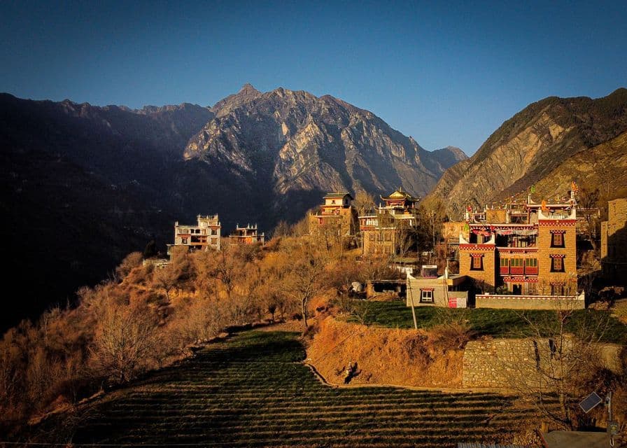 A village with traditional buildings on a sunlit hillside, nestled in a valley with large, rocky mountains in the background.