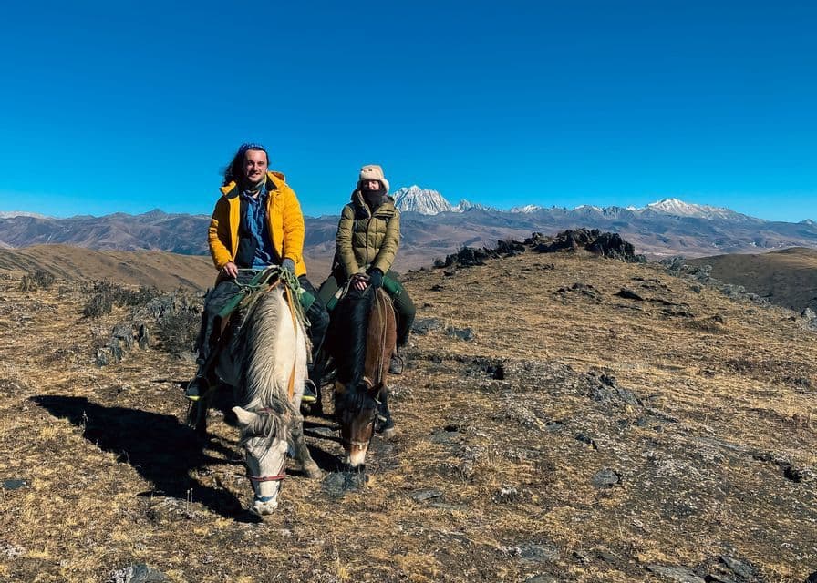 A WeRoad group trip of two people on horseback on a sunny, grassy mountainside with a snow-capped mountain range in the background.