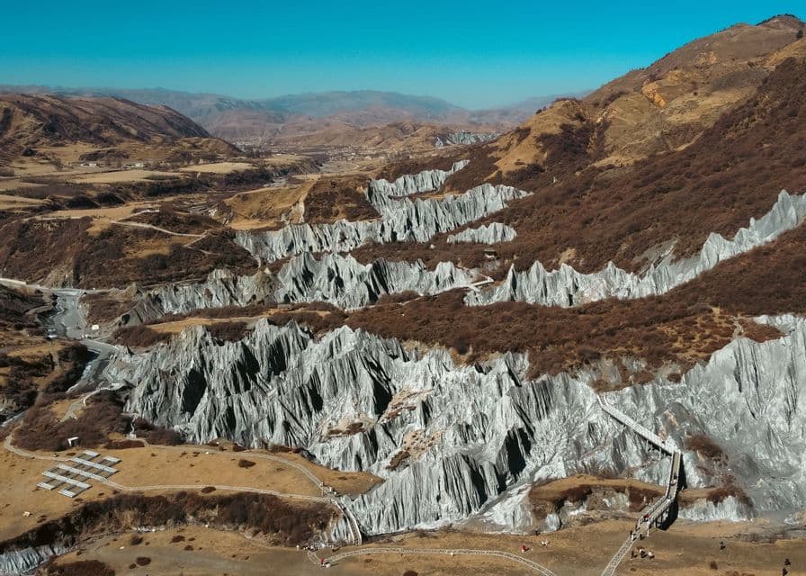 An aerial view of a vast grey stone forest formation with sharp peaks, traversed by winding pathways and a wooden staircase under a clear blue sky.