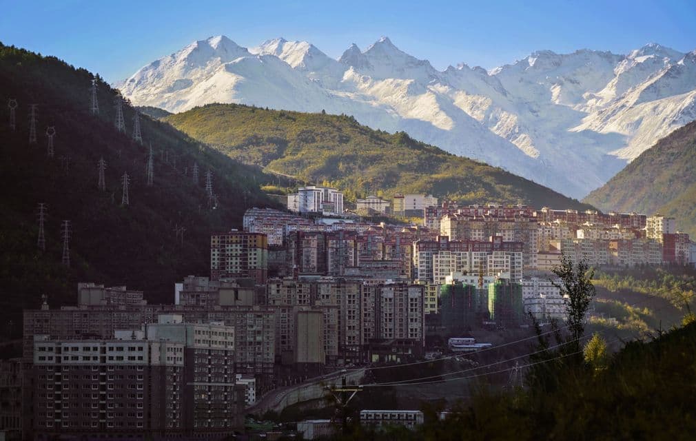 A city of apartment buildings built into a green mountain valley with a large, snow-capped mountain range in the background.