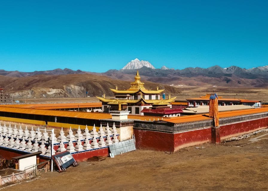 A large monastery with golden roofs and white stupas sits in a valley before arid mountains and a distant snow-capped peak.