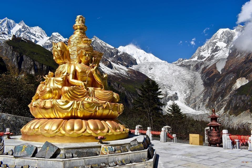 A large golden multi-headed Buddha statue sits on a lotus base, with snow-capped mountains and a glacier in the background.