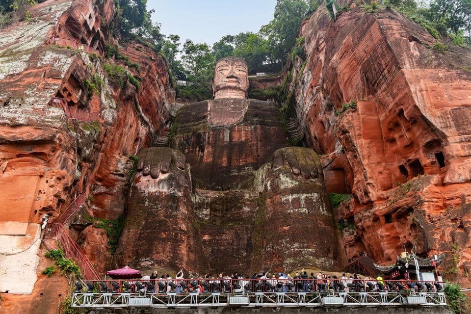 A giant seated Buddha statue is carved into a red sandstone cliff, with tourists on a viewing platform below.