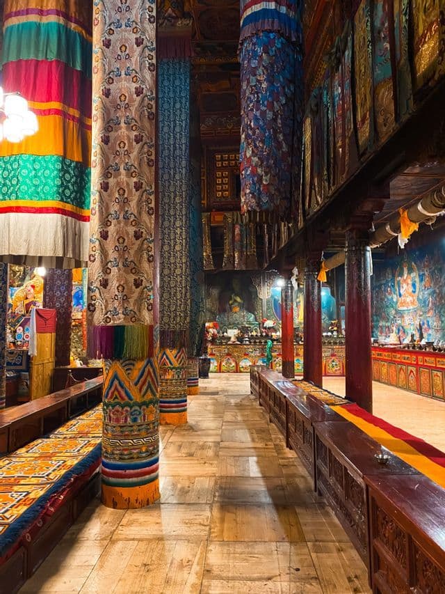 The ornate interior of a temple hall, with pillars wrapped in colorful fabrics and long wooden benches lining a wooden floor.