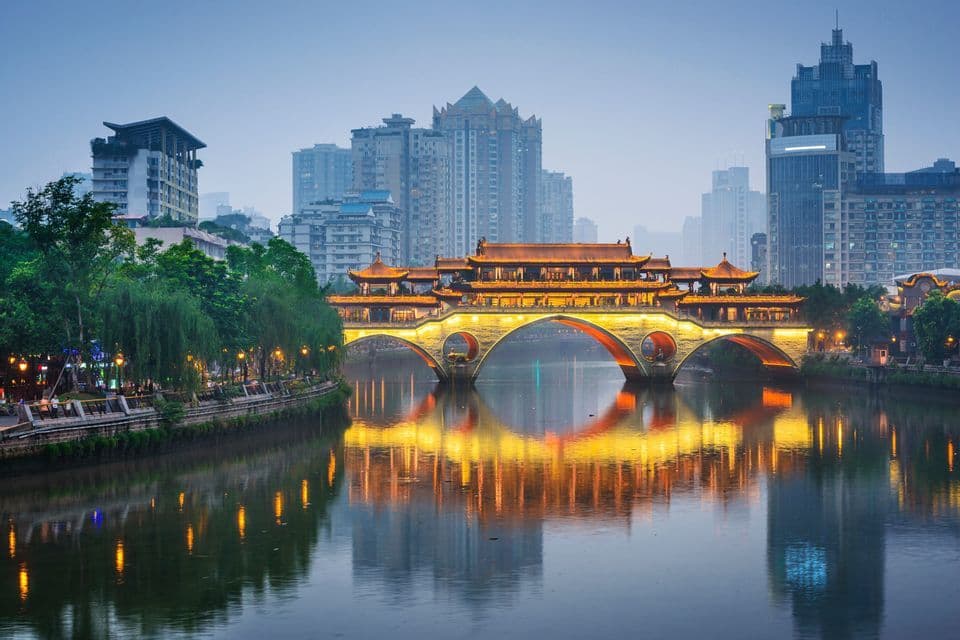 An illuminated, traditional Chinese bridge spans a calm river at dusk, with reflections on the water and a modern city skyline in the background.