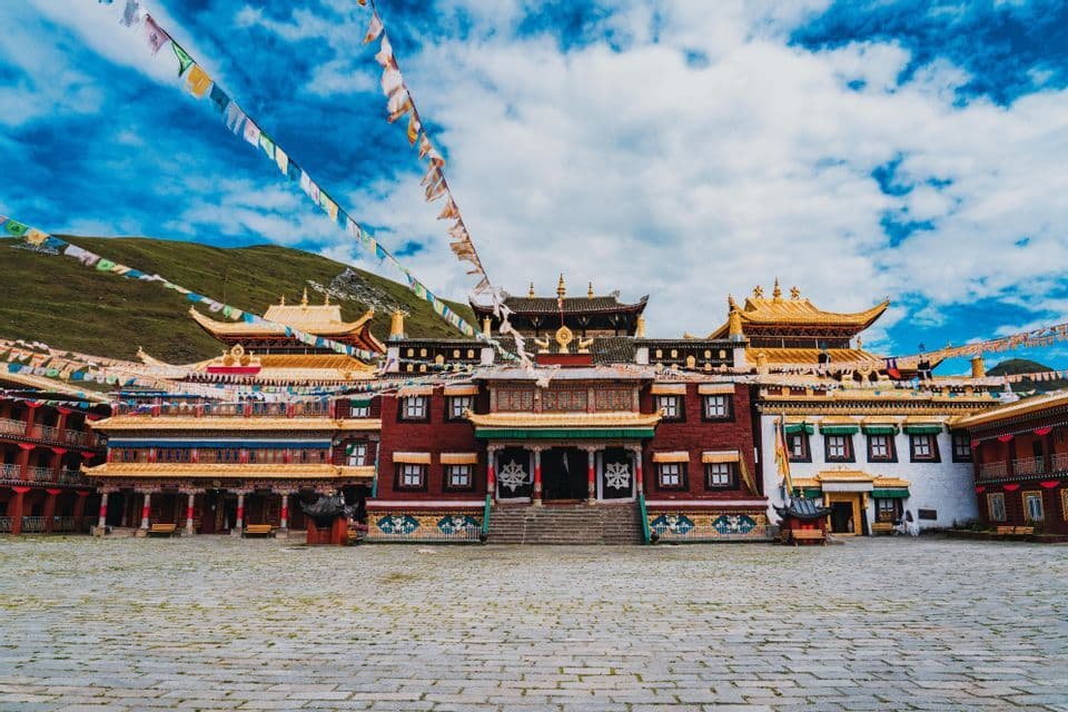 An ornate Buddhist monastery with golden roofs stands across a large stone courtyard, with prayer flags strung above under a blue sky.