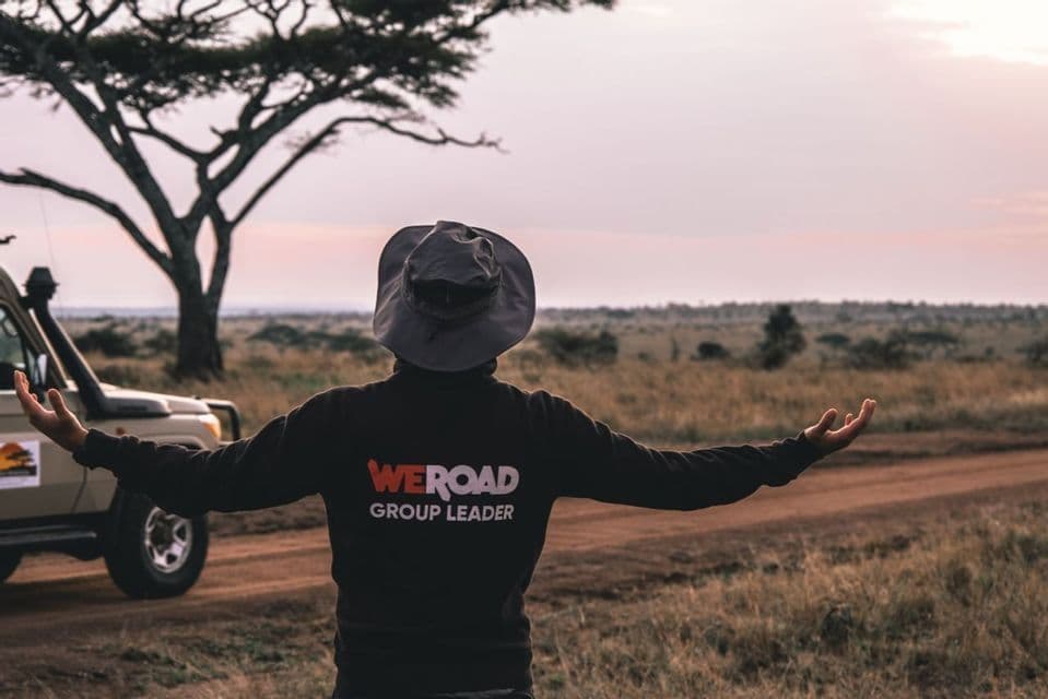 Un capogruppo WeRoad, visto di spalle, sta con le braccia distese in un paesaggio di savana accanto a un veicolo safari al tramonto.