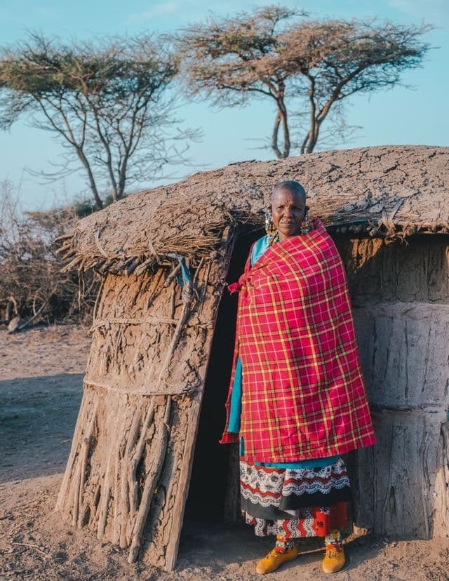 Une femme âgée portant une couverture à carreaux rouges et une tenue traditionnelle se tient à l'entrée d'une hutte en terre.