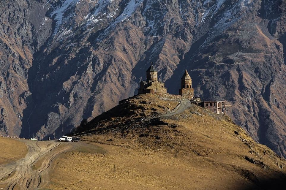 Un monastero in pietra con campanile si erge su una collina erbosa, ai piedi di un'ampia e aspra catena montuosa con chiazze di neve.